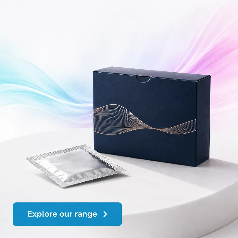 Condom boxes from CondomSales with a metallic wave design on the front, shown beside a small silver foil packet on a white display surface. A blue button reading 'Explore our range >' appears at the bottom left.